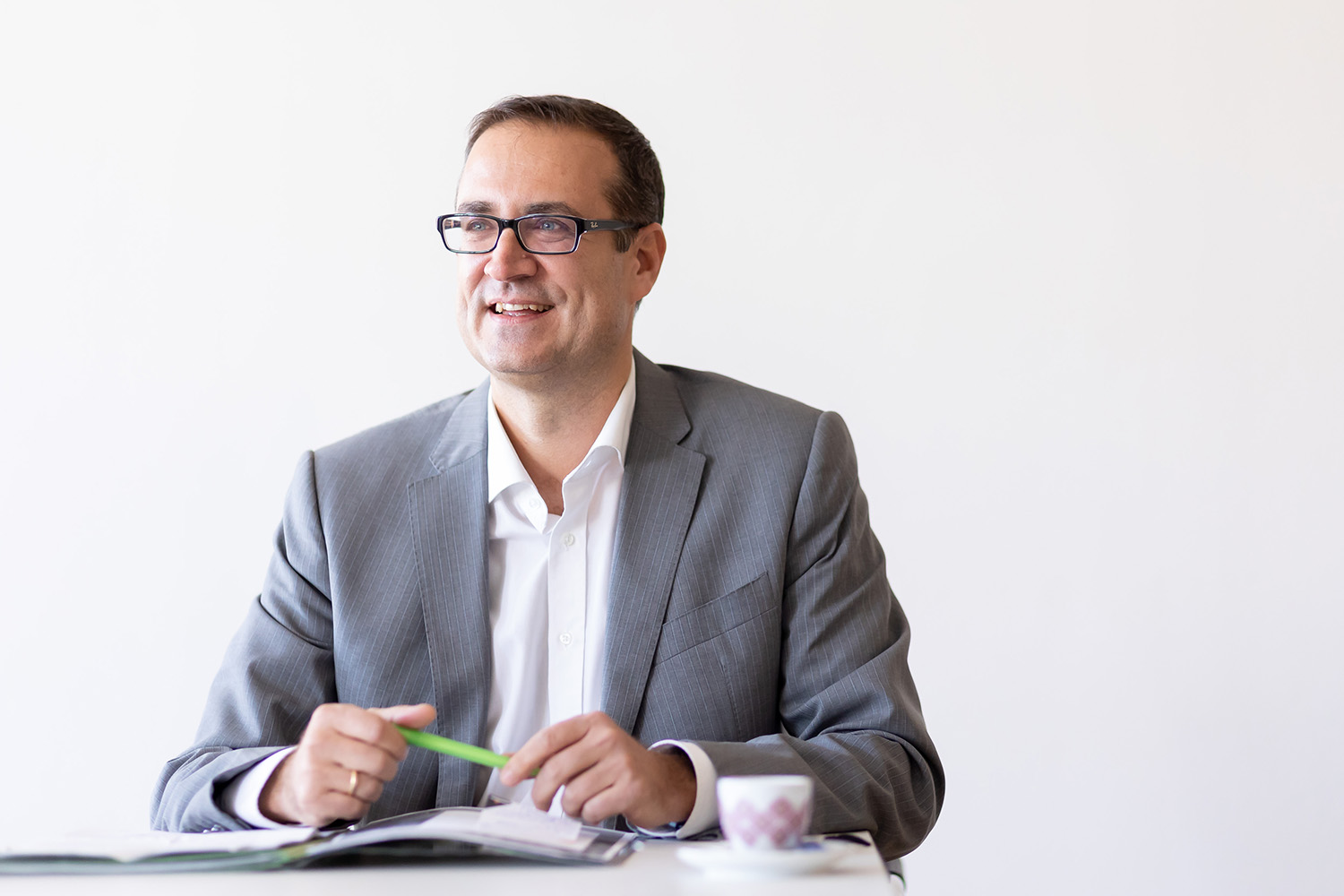 Ein Mann in grauem Anzug und weißem Hemd sitzt an einem Schreibtisch, lächelt und hält einen grünen Stift über ein aufgeschlagenes Notizbuch. Eine kleine Tasse steht auf dem Tisch. Der Hintergrund ist schlicht und weiß.