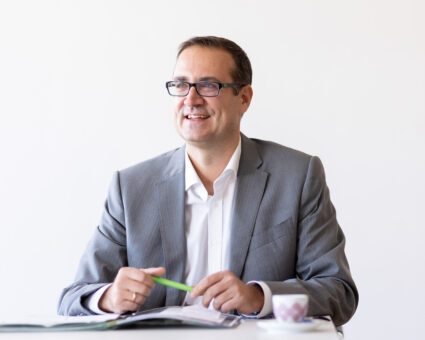 Ein lächelnder Mann in einem grauen Anzug und mit Brille sitzt an einem Schreibtisch mit einem aufgeschlagenen Notizbuch und einem grünen Stift in der Hand. Eine kleine rosa-weiße Tasse steht auf dem Tisch. Der Hintergrund ist schlicht und weiß.