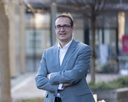 Ein Mann mit Brille und hellgrauem Anzug steht im Freien, verschränkt die Arme und lächelt. Der Hintergrund ist unscharf und zeigt Bäume, Gebäude und Fahrräder.