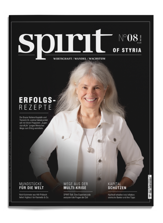 Eine lächelnde Frau mit langen weißen Haaren, die eine weiße Jacke trägt, steht vor einem schwarzen Hintergrund auf dem Cover des Magazins "spirit", Ausgabe 8.