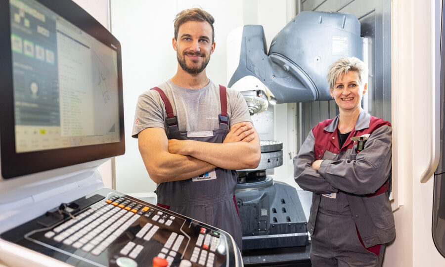 Zwei Fabrikarbeiter in Overalls stehen lächelnd und mit verschränkten Armen vor Industriemaschinen und einem Computerbedienfeld in einer modernen Produktionsstätte.