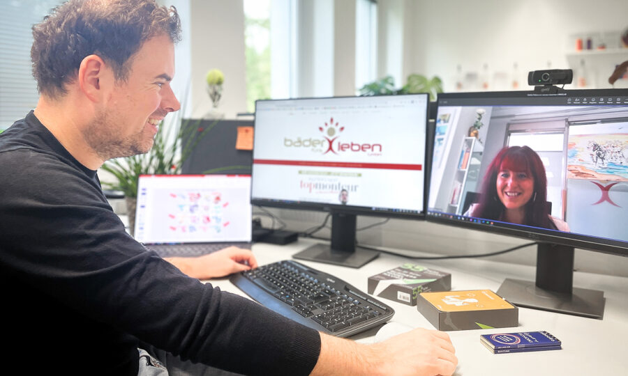 Ein Mann sitzt an einem Schreibtisch und arbeitet an einem Computer mit zwei Monitoren, während er auf einem Bildschirm einen Videochat mit einer Frau führt. Auf dem Schreibtisch liegen Büromaterial und elektronische Geräte, im Hintergrund sind Fenster und Regale zu sehen.