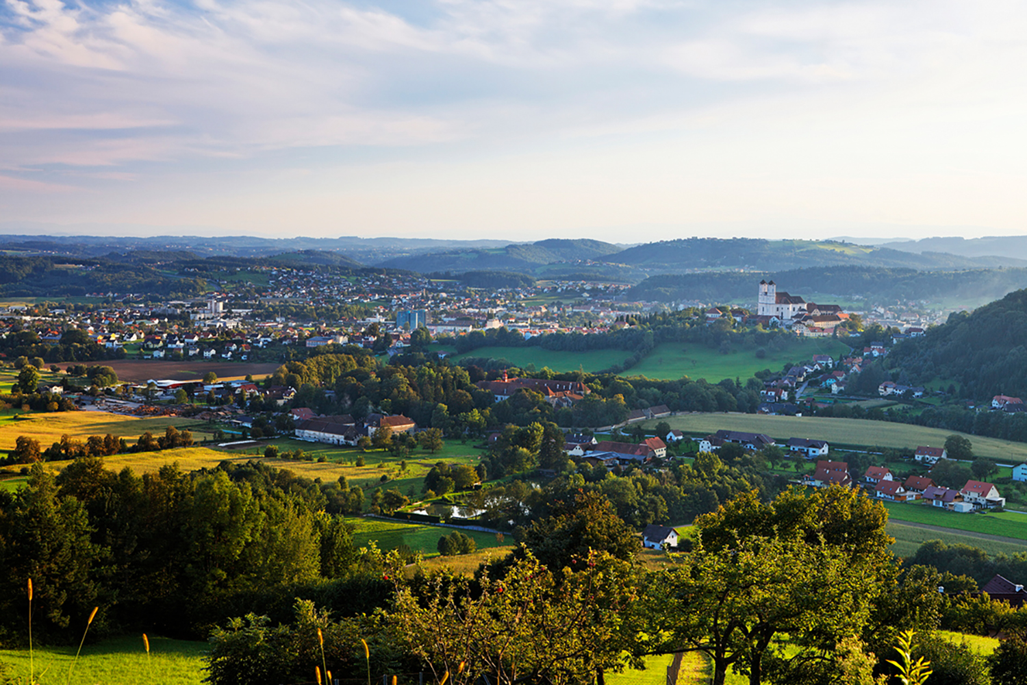 Ein malerischer Blick auf eine kleine Stadt mit verstreuten Häusern, grünen Feldern und Bäumen unter einem teilweise bewölkten Himmel. Eine große weiße Kirche oder ein Schloss ist auf einem Hügel in der Ferne zu sehen, umgeben von sanften Hügeln und Wäldern.