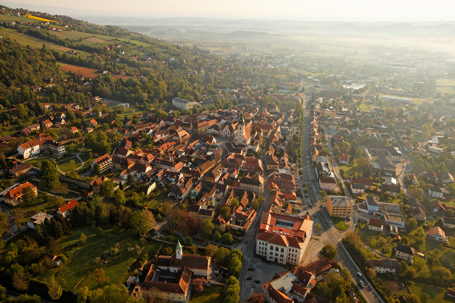 Luftaufnahme einer europäischen Kleinstadt mit roten Dächern, verwinkelten Straßen und üppig grünen Hügeln im Hintergrund im dunstigen, goldenen Morgenlicht.