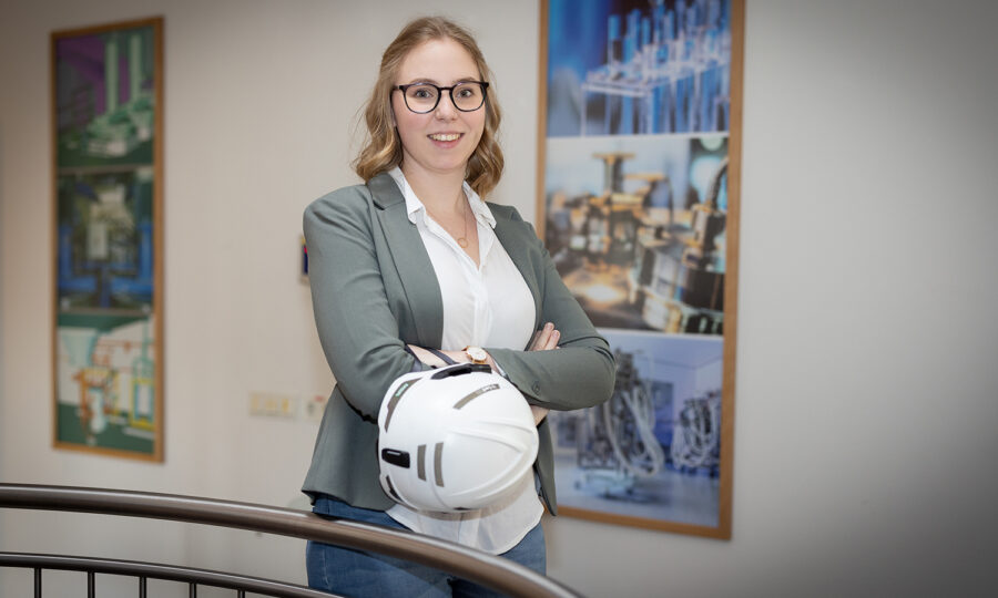 Eine Frau mit Brille und schulterlangem Haar steht in einem Innenraum, lächelt mit verschränkten Armen und hält einen weißen Schutzhelm. Sie trägt einen Blazer und steht vor zwei bunten Plakaten mit wissenschaftlichen Themen.