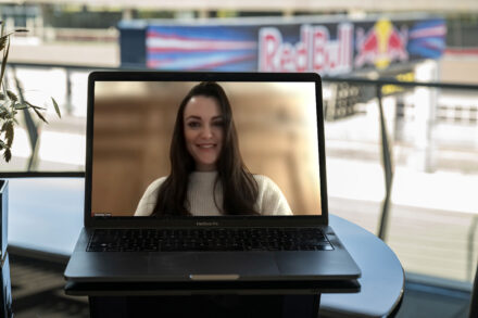 Ein Laptop auf einem Tisch zeigt ein Videogespräch mit einer lächelnden Frau in einem weißen Pullover. Im verschwommenen Hintergrund sind ein Red Bull-Schild und eine Rennstrecke zu sehen.
