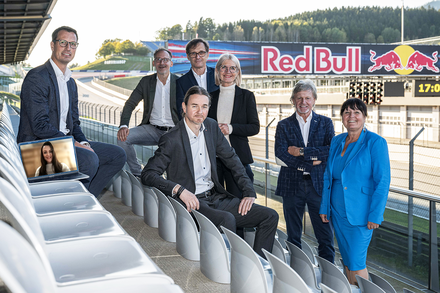 Sieben Personen in Geschäftskleidung posieren lächelnd auf Stadionsitzen; ein Laptop auf einem Sitz zeigt eine Frau bei einem Videogespräch. Im Hintergrund sind die Rennstrecke und das Logo des Red Bull Rings zu sehen.