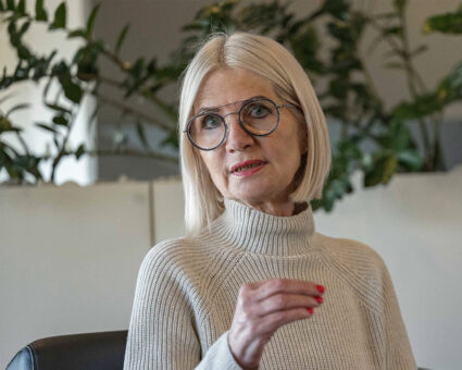 Eine ältere Frau mit glattem weißem Haar und Brille, die einen hellen Strickpullover trägt, gestikuliert mit ihrer Hand, während sie spricht. Im Hintergrund sind Grünpflanzen zu sehen.