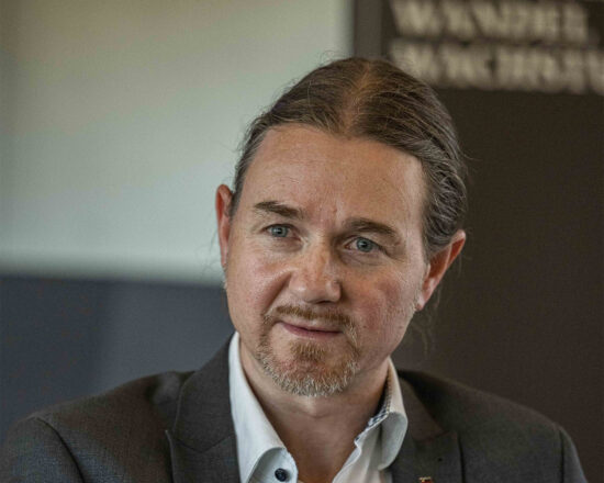 Ein Mann mit langen braunen Haaren, die nach hinten gebunden sind, einem kurzen Bart und Schnurrbart, der einen grauen Blazer und ein weißes Hemd trägt, schaut in einem gut beleuchteten Innenraum leicht zur Seite.