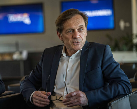 Ein Mann in einem dunklen Anzug und weißem Hemd sitzt in einem Haus und hält eine Brille in den Händen. Im Hintergrund sind zwei verschwommene Bildschirme zu sehen. Er scheint in ein Gespräch vertieft zu sein.