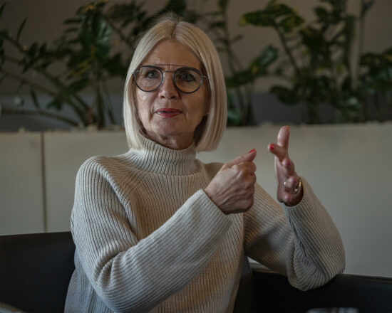 Eine ältere Frau mit glattem weißem Haar, die eine Brille und einen hellen Strickpullover trägt, sitzt in einem Haus und spricht mit ihren Händen. Im Hintergrund sind Grünpflanzen zu sehen.