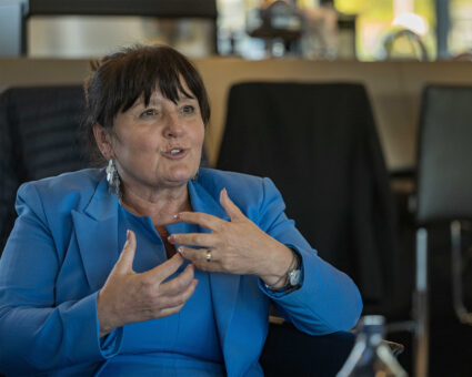Eine Frau mit dunklem Haar in einem blauen Blazer gestikuliert mit ihren Händen, während sie spricht. Sie sitzt in einem Innenraum mit unscharfen Stühlen und einem Tisch im Hintergrund.