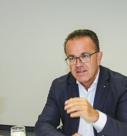 Ein Mann in einem dunklen Blazer und mit Brille spricht angeregt während eines Gesprächs an einem Tisch und gestikuliert mit der Hand. Im Vordergrund ist eine weitere Person mit grauem Haar und Brille zu sehen, die ihm gegenübersteht.