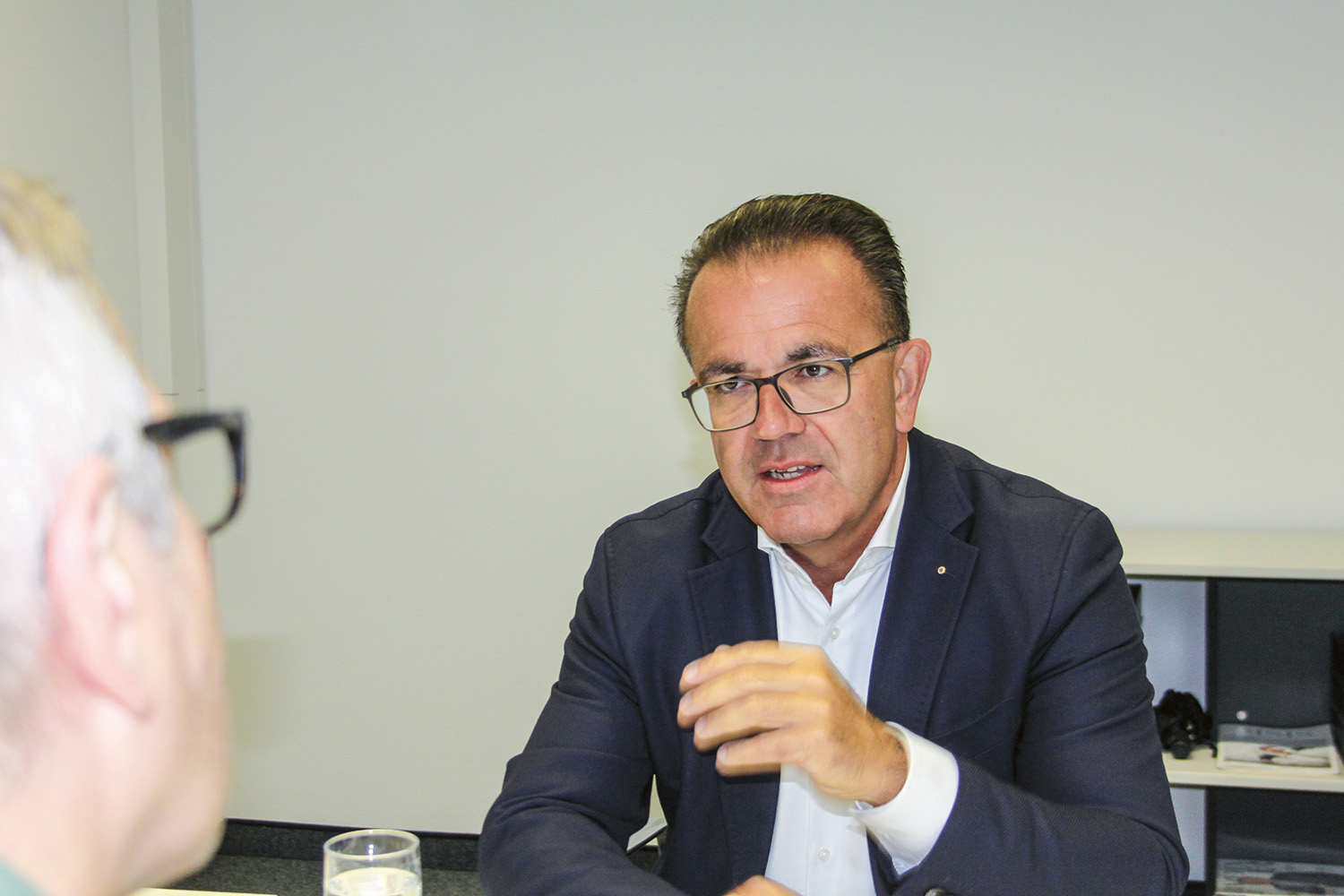 Ein Mann in einem dunklen Blazer und mit Brille spricht angeregt während eines Gesprächs an einem Tisch und gestikuliert mit der Hand. Im Vordergrund ist eine weitere Person mit grauem Haar und Brille zu sehen, die ihm gegenübersteht.