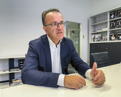 Ein Mann im Anzug und mit Brille sitzt an einem weißen Schreibtisch in einem modernen Büro und gestikuliert mit seiner Hand, während er spricht. Im Hintergrund sind Regale mit Zeitschriften und Aktenordnern zu sehen.