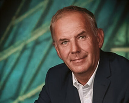 Ein Mann mittleren Alters mit kurzen grauen Haaren, der eine dunkle Anzugsjacke und ein weißes Hemd trägt, steht leicht lächelnd vor einem blau-grün gemusterten Hintergrund.