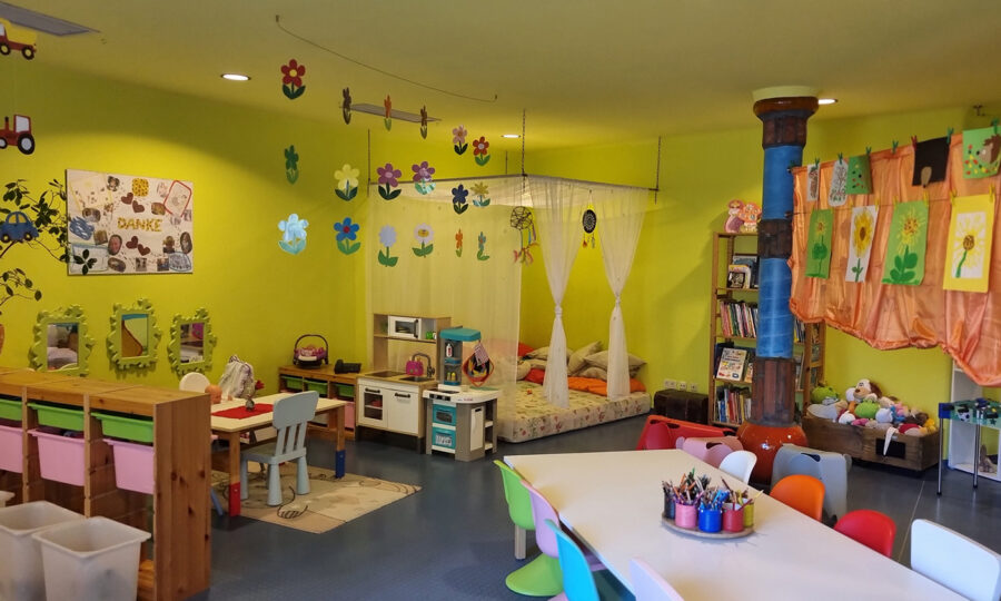 Ein farbenfrohes Klassenzimmer mit kleinen Tischen und Stühlen, Malutensilien, Kinderzeichnungen an den Wänden, einer Spielküche, Bücherregalen, einer gemütlichen Bettecke mit Kissen und verspielten Dekorationen an der Decke.