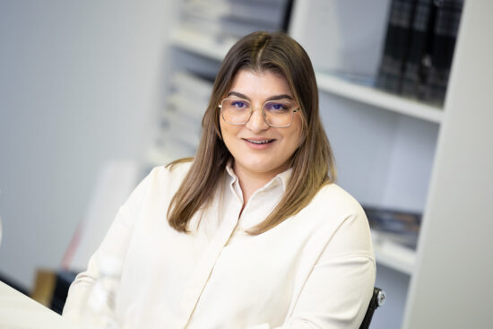 Eine Person mit langen braunen Haaren und Brille lächelt, während sie an einem Schreibtisch in einem Büro sitzt. Sie trägt ein helles Hemd, und im Hintergrund sind Regale mit Büchern und Akten zu sehen.