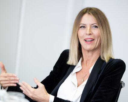 Eine Frau mit glattem, schulterlangem blondem Haar lächelt und gestikuliert mit ihren Händen, während sie spricht. Sie trägt einen schwarzen Blazer über einer weißen Bluse und sitzt in einer hellen, modernen Umgebung.