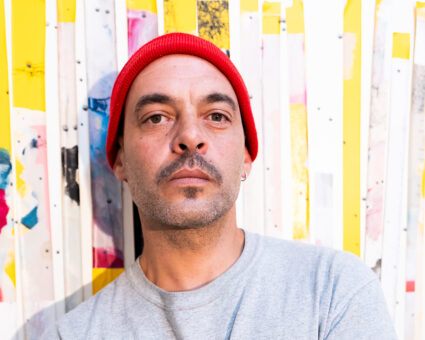 Ein Mann mit roter Mütze und grauem T-Shirt steht vor einem bunten, abstrakten Hintergrund mit gelben und weißen Streifen und blickt mit neutralem Blick direkt in die Kamera.