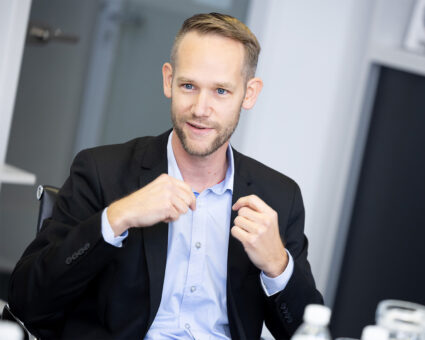 Ein Mann im Anzug sitzt an einem Tisch und gestikuliert mit beiden Händen, als würde er etwas erklären. Er hat kurzes Haar und einen gestutzten Bart und scheint sich in einer Büroumgebung zu unterhalten.