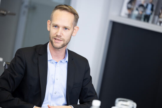 Ein Mann in einem schwarzen Anzug und hellblauem Hemd sitzt an einem Schreibtisch und führt ein Gespräch. Der Hintergrund ist eine moderne Büroumgebung mit verschwommenen Regalen und einer Tür.