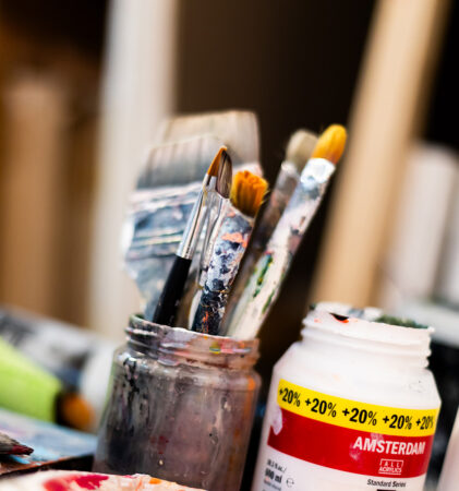 Eine Nahaufnahme von Pinseln in einem Glasgefäß neben einem offenen Behälter mit Amsterdamer Acrylfarbe und einer weißen Flasche, alles auf einem unordentlichen Künstlerarbeitsplatz.