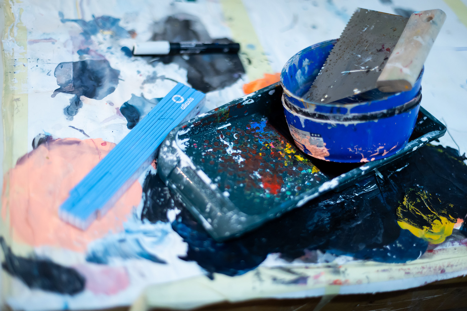 Ein unordentlicher Arbeitsbereich eines Künstlers mit einem blauen Farbtablett, einem Pinsel, einer blauen Schale, einem Metallspachtel, einem Lineal und verschiedenen Farbspritzern auf einer weißen Fläche.