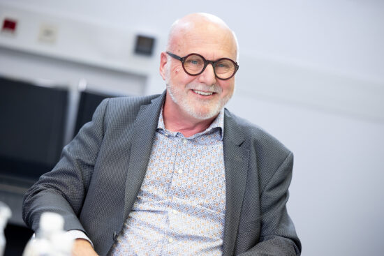 Ein lächelnder älterer Mann mit Brille, einer Glatze und einem grauen Bart sitzt in einem Büro. Er trägt ein gemustertes Hemd und einen grauen Blazer. Der Hintergrund ist unscharf.
