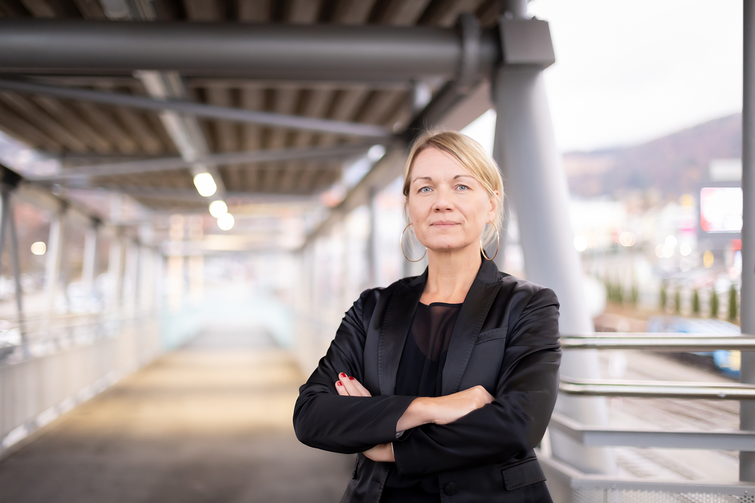 Eine selbstbewusste Frau in einem schwarzen Anzug steht mit verschränkten Armen auf einer modernen Fußgängerbrücke und blickt direkt in die Kamera, während die Stadtkulisse im Hintergrund verschwimmt.