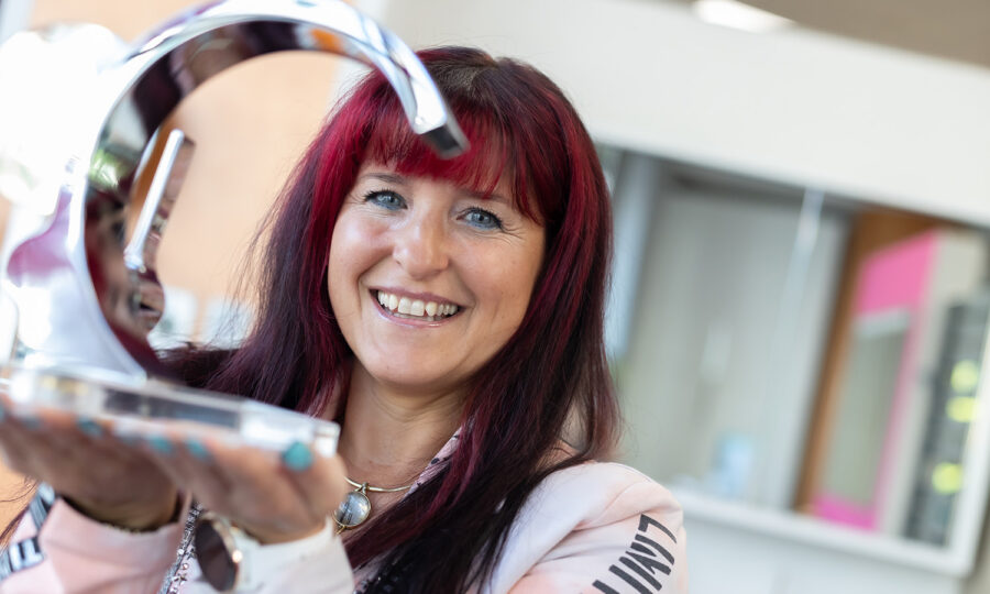 Eine Frau mit langen dunklen Haaren und roten Strähnen lächelt, während sie eine glänzende, abstrakte Metalltrophäe in beiden Händen hält. Sie befindet sich in einem Innenraum, trägt eine helle Jacke, und der Hintergrund zeigt ein verschwommenes modernes Interieur.