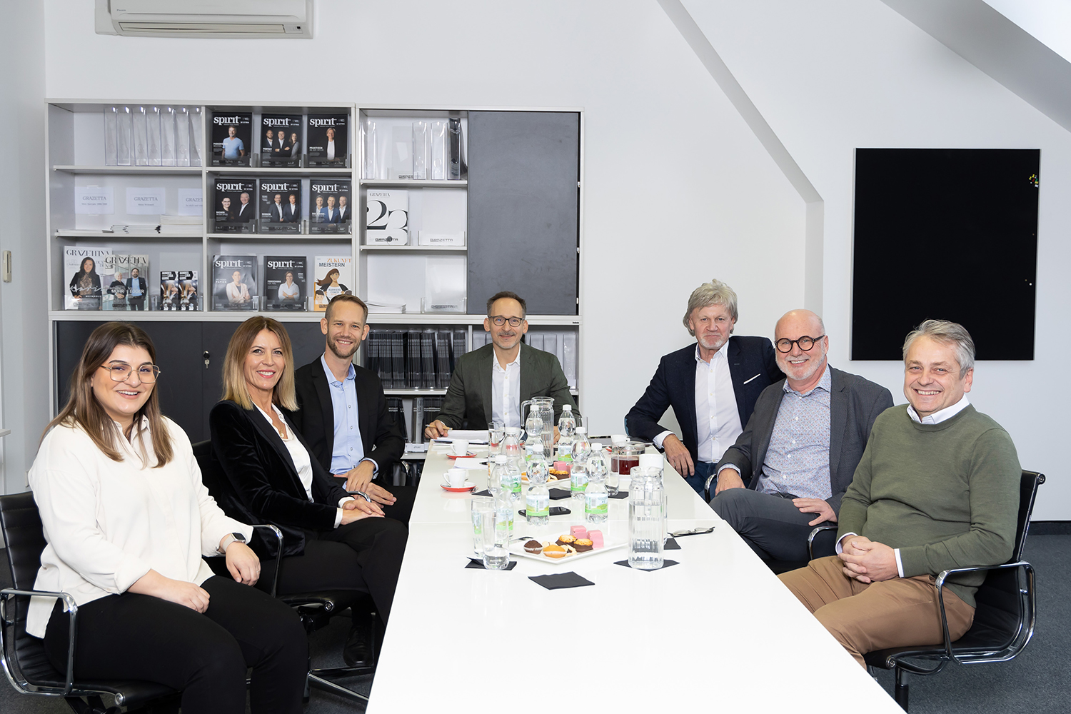 Sieben professionell gekleidete Personen sitzen um einen Konferenztisch mit Papieren, Getränken und Snacks und lächeln in die Kamera in einem modernen Bürobesprechungsraum mit Zeitschriften in Regalen im Hintergrund.