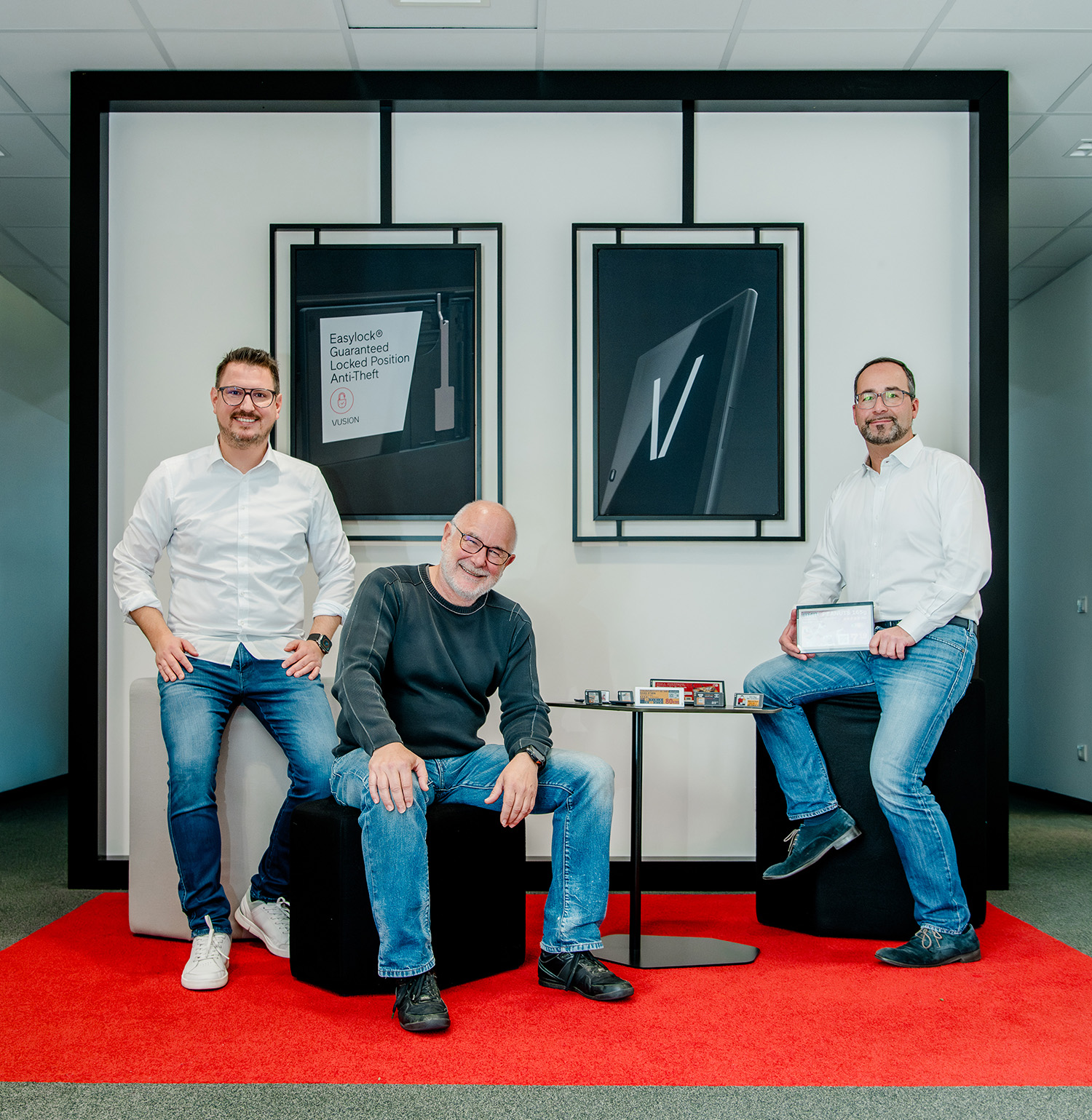 Drei Männer in Freizeitkleidung posieren in einem modernen Büroraum mit weißen Wänden und rotem Teppich. Zwei von ihnen stehen, einer sitzt. Hinter ihnen hängen zwei gerahmte Poster mit technischen Produkten an der Wand.