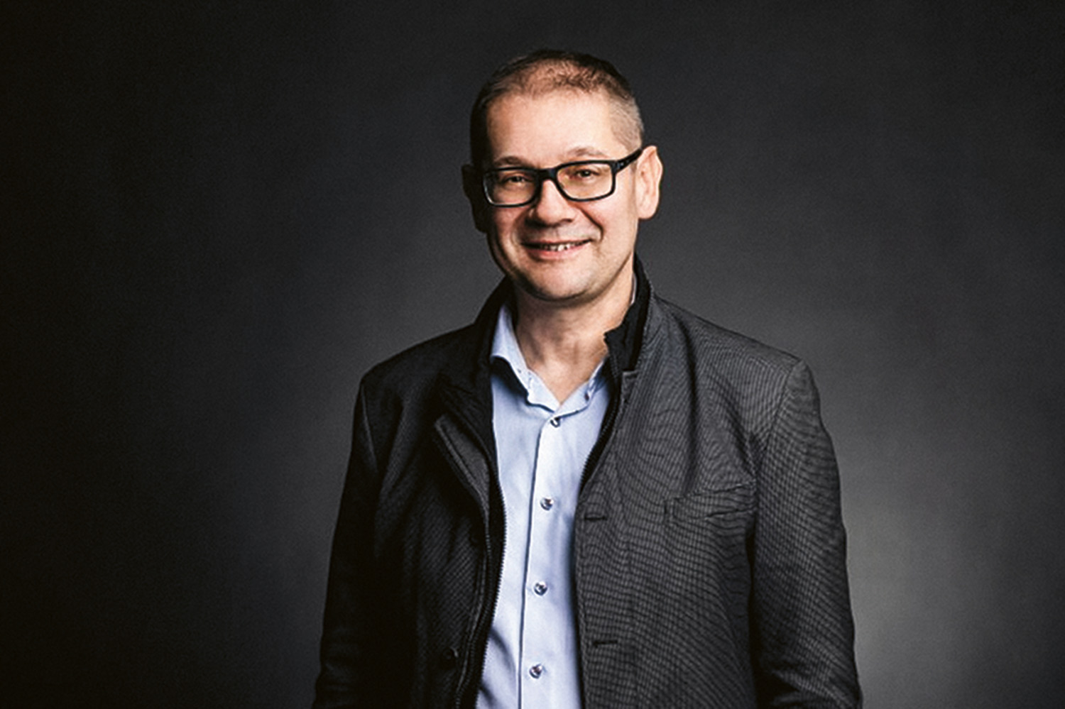 Ein Mann mit kurzen Haaren und Brille, der ein hellblaues Hemd und einen dunklen strukturierten Blazer trägt, lächelt, während er vor einem schlichten dunklen Hintergrund steht.