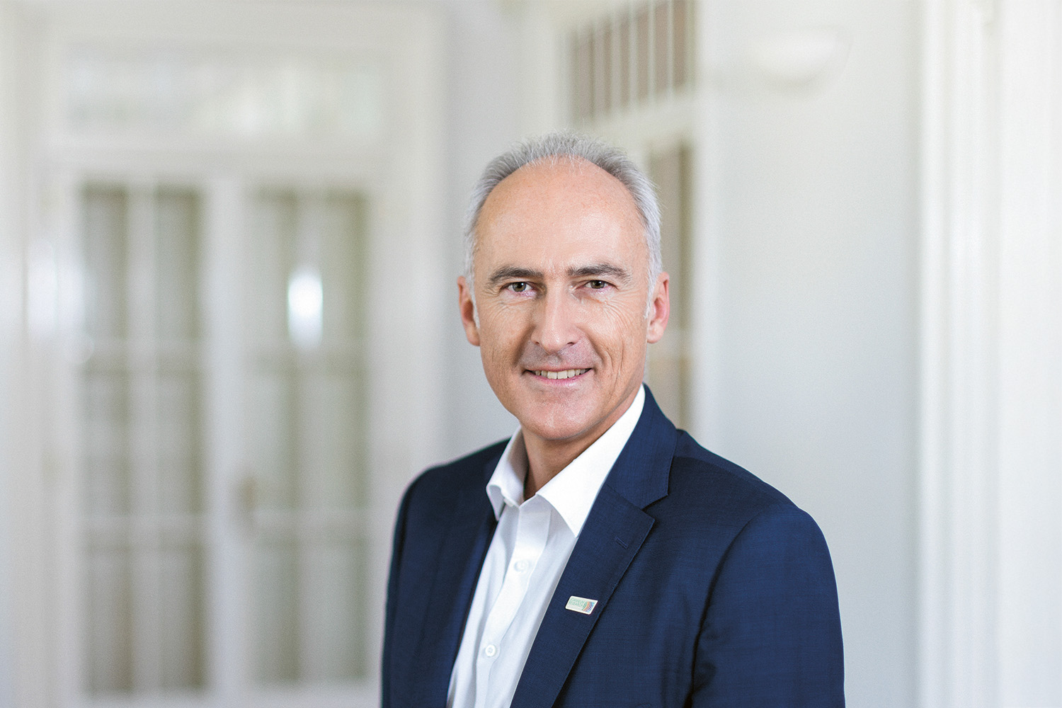 Ein Mann mittleren Alters mit kurzen grauen Haaren, der ein weißes Hemd und eine dunkelblaue Anzugsjacke trägt, steht lächelnd vor einer gläsernen Doppeltür.