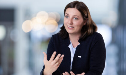 Eine Frau mit schulterlangem braunem Haar, die eine dunkle Jacke über einem weißen Oberteil trägt, gestikuliert mit ihren Händen, während sie spricht. Der Hintergrund ist mit hellen Lichtern sanft verschwommen.