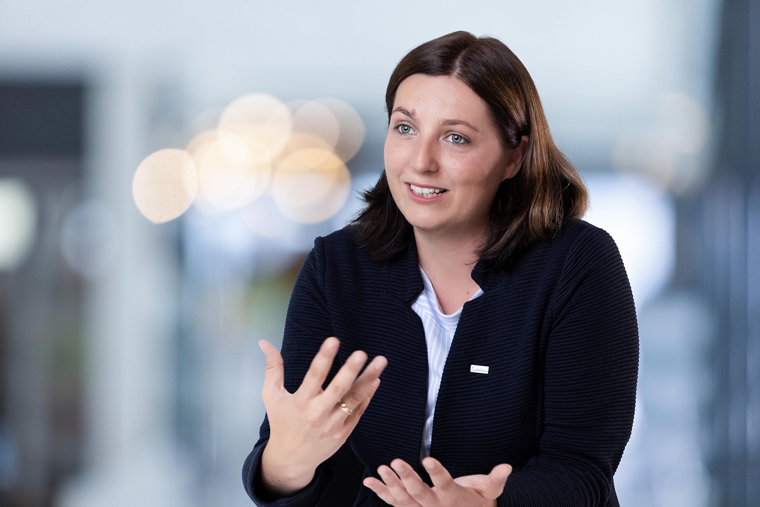 Eine Frau mit schulterlangem braunem Haar, die eine dunkle Jacke über einem weißen Oberteil trägt, gestikuliert mit ihren Händen, während sie spricht. Der Hintergrund ist mit hellen Lichtern sanft verschwommen.