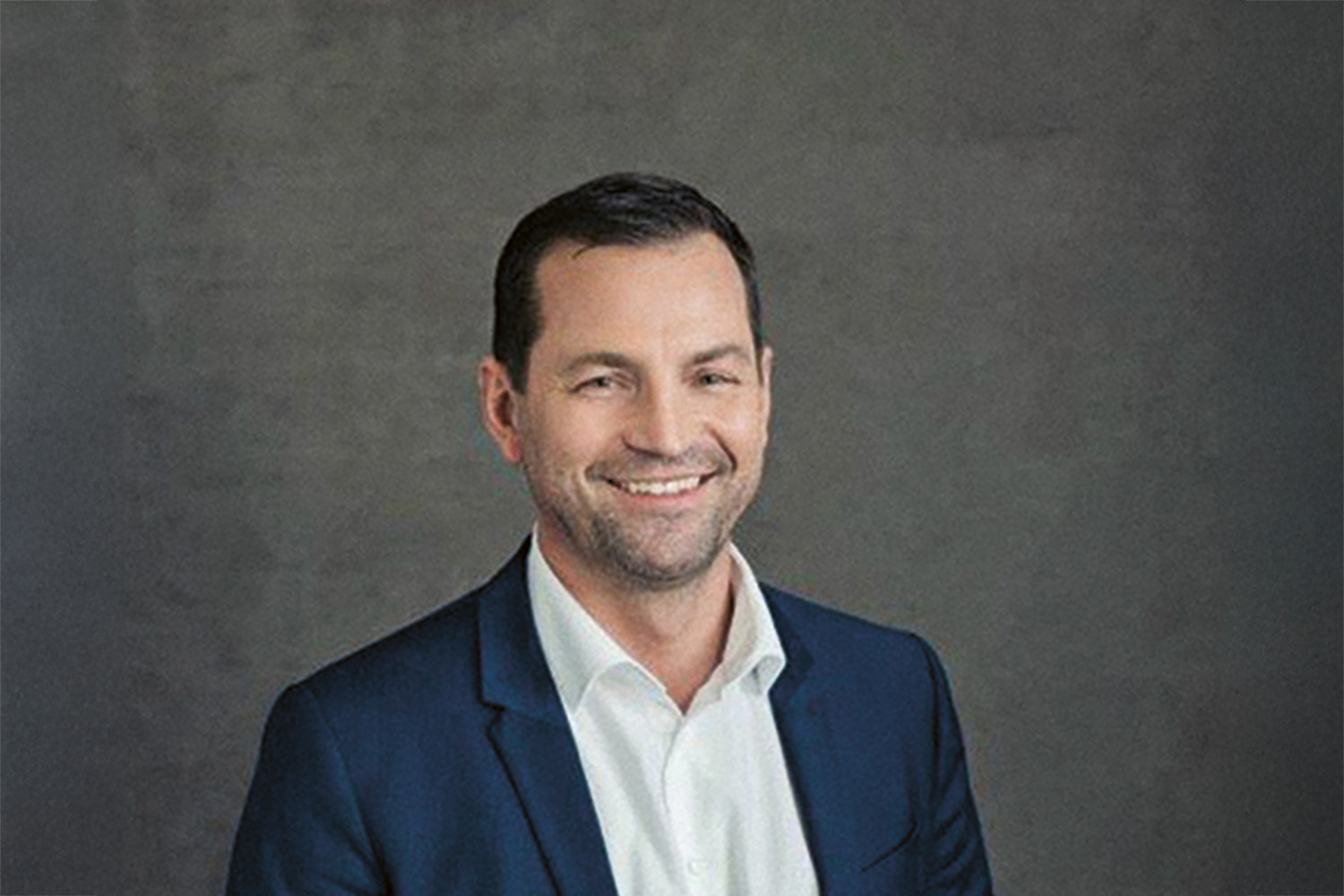 Ein Mann in einem dunkelblauen Anzug und weißem Hemd lächelt vor einem schlichten, grauen Hintergrund. Er hat kurzes dunkles Haar und einen gestutzten Bart.