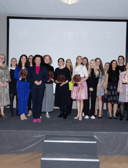 Eine Gruppe von Frauen und einige Männer stehen auf einer Bühne, einige halten Auszeichnungen oder Blumensträuße in der Hand. Sie tragen formelle und halbformelle Kleidung und posieren für ein Gruppenfoto bei einer Indoor-Veranstaltung.