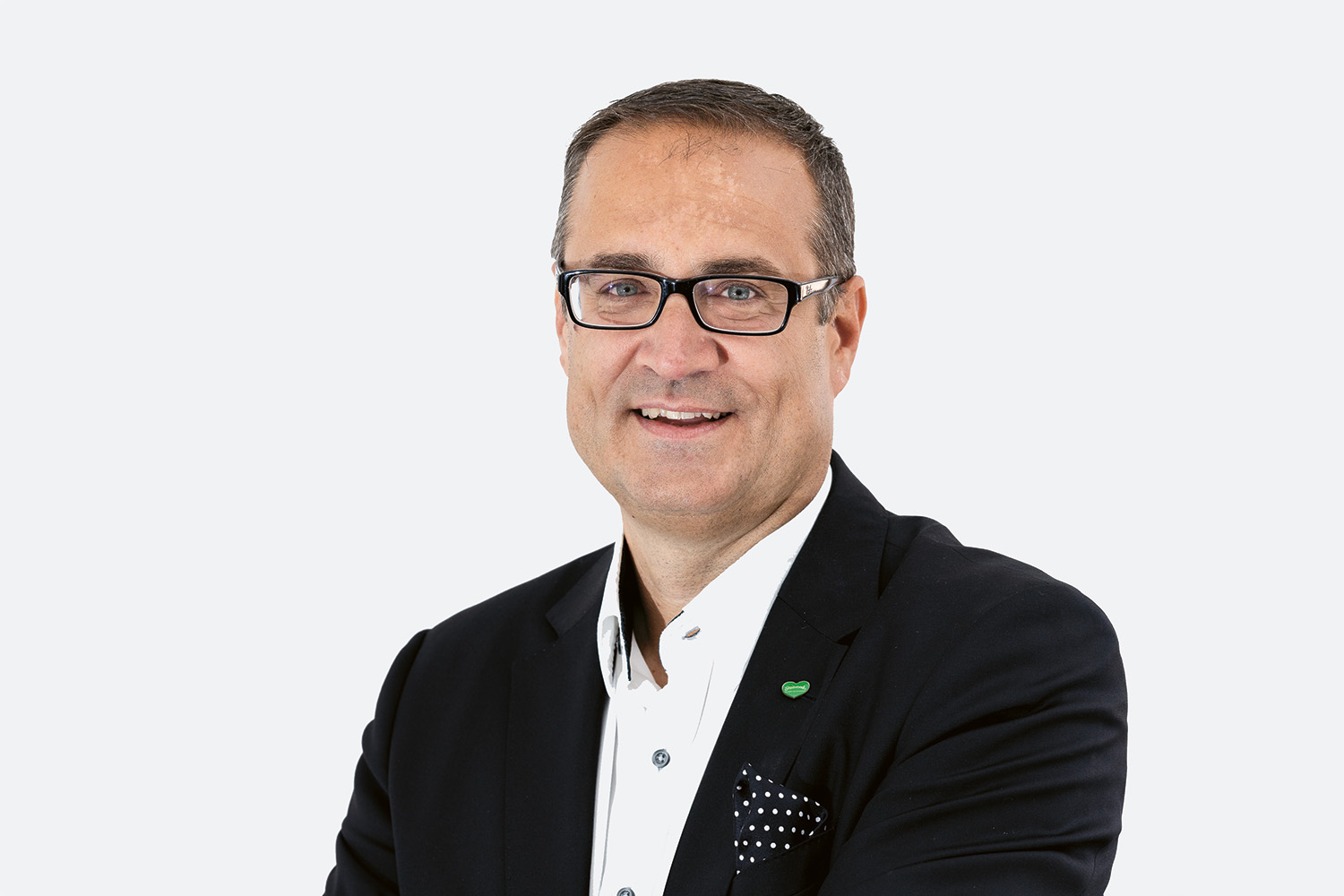 Ein Mann, der eine Brille, einen dunklen Blazer und ein weißes Hemd trägt, lächelt in die Kamera vor einem schlichten, hellen Hintergrund. Er hat kurzes braunes Haar und ein gemustertes Einstecktuch in seiner Jacke.