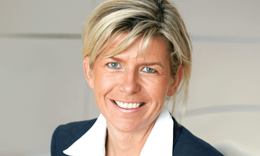 Eine Frau mit blondem Haar in einem professionellen dunklen Blazer und weißem Hemd mit Kragen lächelt in die Kamera vor einem neutralen Hintergrund.