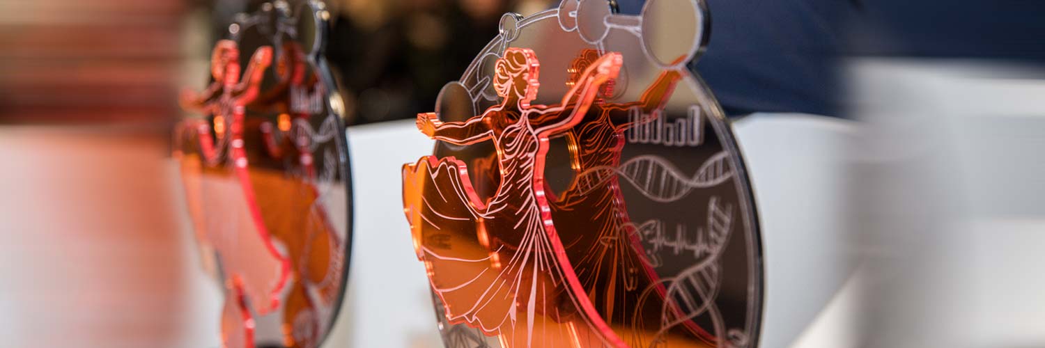 Nahaufnahme von zwei durchscheinenden orangefarbenen Acrylpreisen mit einem eingravierten Bild von tanzenden Paaren und wissenschaftlichen Symbolen, die auf einem Tisch mit unscharfem Hintergrund stehen.