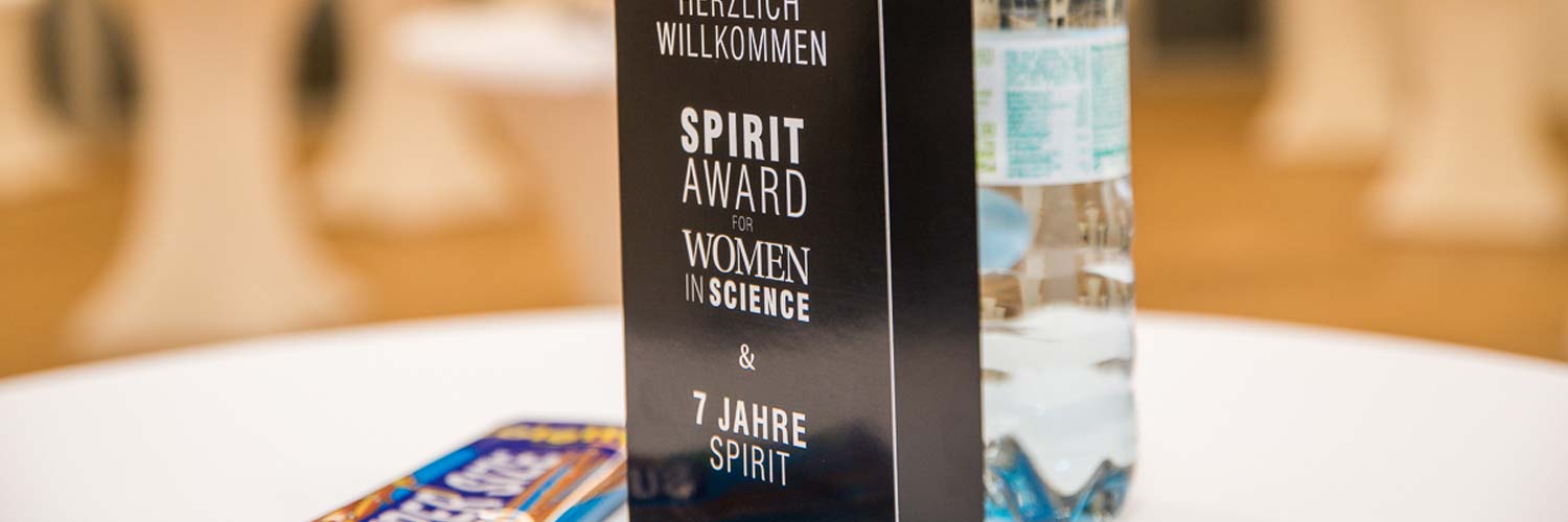 Auf einer schwarzen Preistafel auf einem weißen Tisch steht "Spirit Award for Women in Science & 7 Jahre Spirit", daneben eine Flasche Wasser und eine teilweise sichtbare Tafel Schokolade.