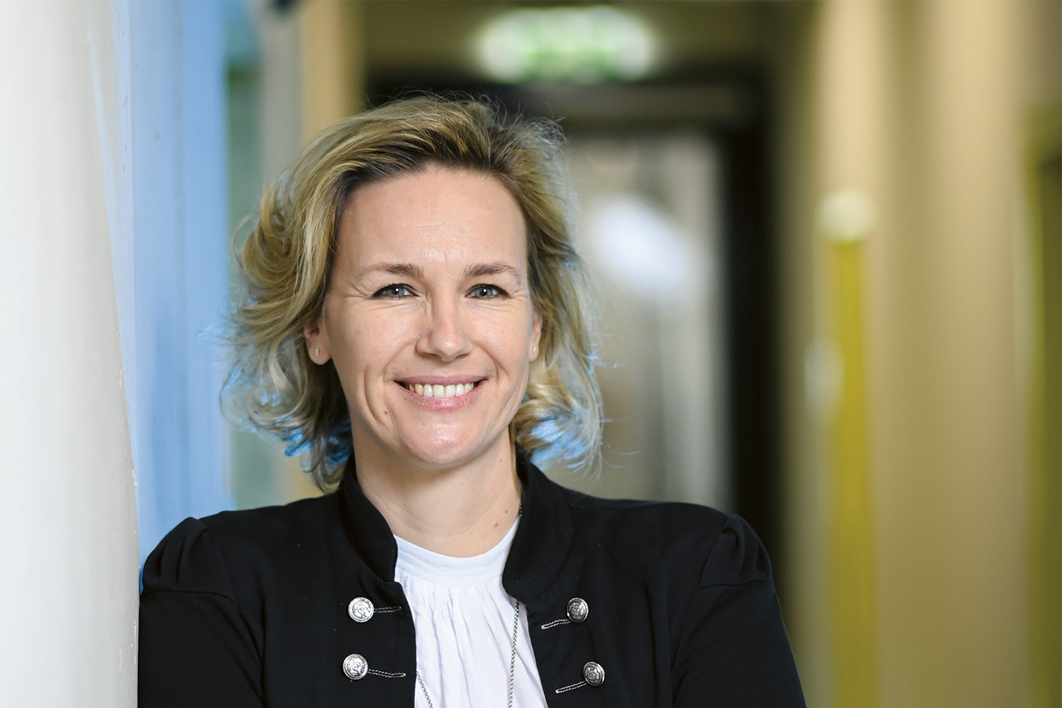 Eine Frau mit gewelltem blondem Haar, die eine schwarze Jacke mit Zierknöpfen und ein weißes Oberteil trägt, steht in einem Haus und lächelt in die Kamera. Der Hintergrund ist leicht unscharf und zeigt einen Hausflur.