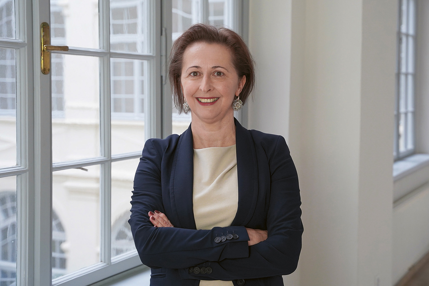 Eine Frau mit kurzen braunen Haaren, die einen dunklen Blazer über einem hellen Oberteil trägt, steht in einem Haus mit verschränkten Armen und lächelt in die Kamera. Im Hintergrund sind große Fenster und weiße Wände zu sehen.