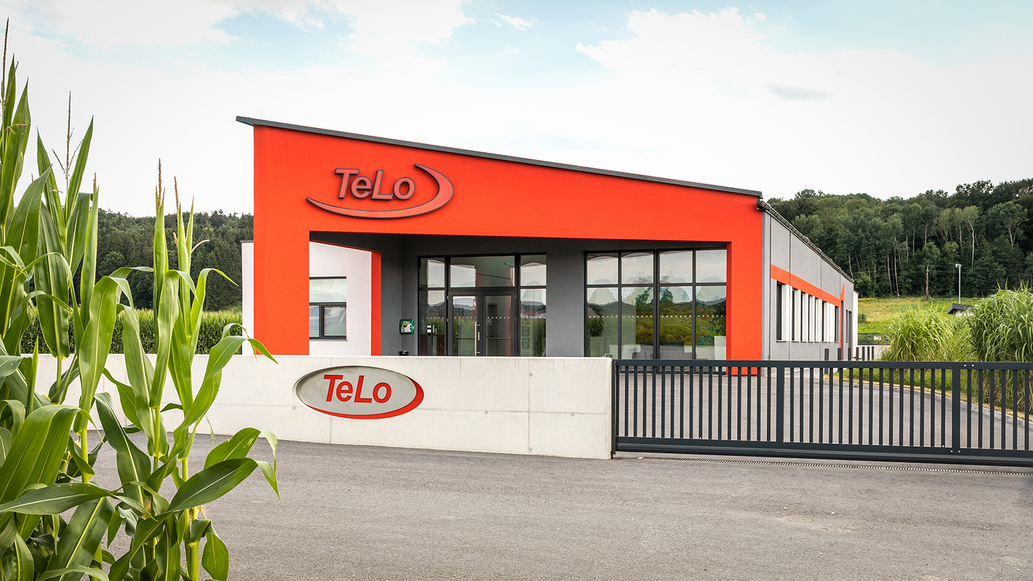 Ein modernes Gebäude mit leuchtend roten Akzenten und großen Fenstern mit der Aufschrift "TeLo" befindet sich hinter einem Betonzaun und einem schwarzen Tor. Im Vordergrund sind Maispflanzen zu sehen, und im Hintergrund stehen Bäume.