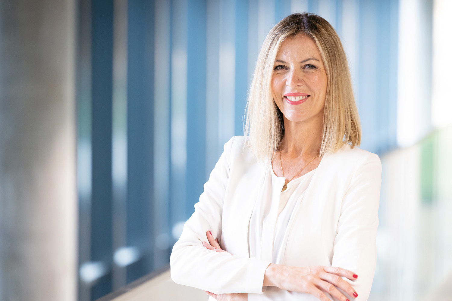 Eine Frau mit glattem blondem Haar, die einen weißen Blazer trägt, steht mit verschränkten Armen im Haus und lächelt selbstbewusst. Im Hintergrund sind blaue Tafeln und ein verschwommener Korridor zu sehen.