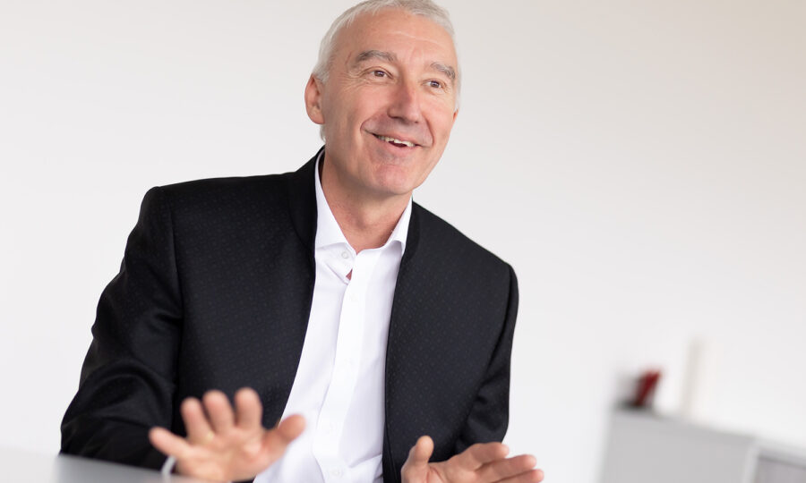 Ein lächelnder älterer Mann mit kurzen grauen Haaren, der eine dunkle Anzugsjacke und ein weißes Hemd trägt, gestikuliert mit den Händen, während er an einem Tisch in einer hellen, minimalistischen Büroumgebung sitzt.