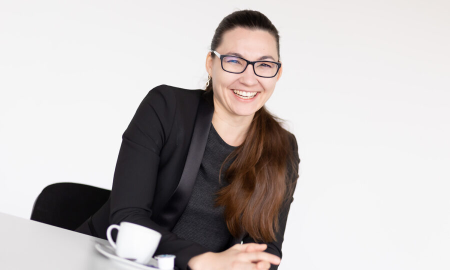 Eine Frau mit langen braunen Haaren und Brille, die einen schwarzen Blazer trägt, sitzt an einem weißen Tisch und lächelt. Eine weiße Kaffeetasse und eine Untertasse stehen auf dem Tisch vor ihr. Der Hintergrund ist einfarbig weiß.