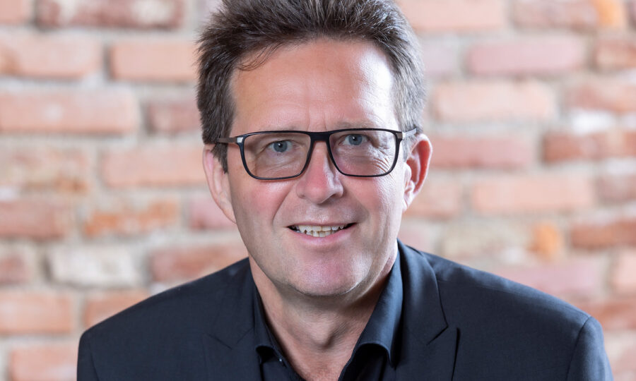 Ein Mann mittleren Alters mit kurzen braunen Haaren, der eine Brille und eine dunkle Anzugsjacke trägt, lächelt vor einem unscharfen Backsteinmauer-Hintergrund.